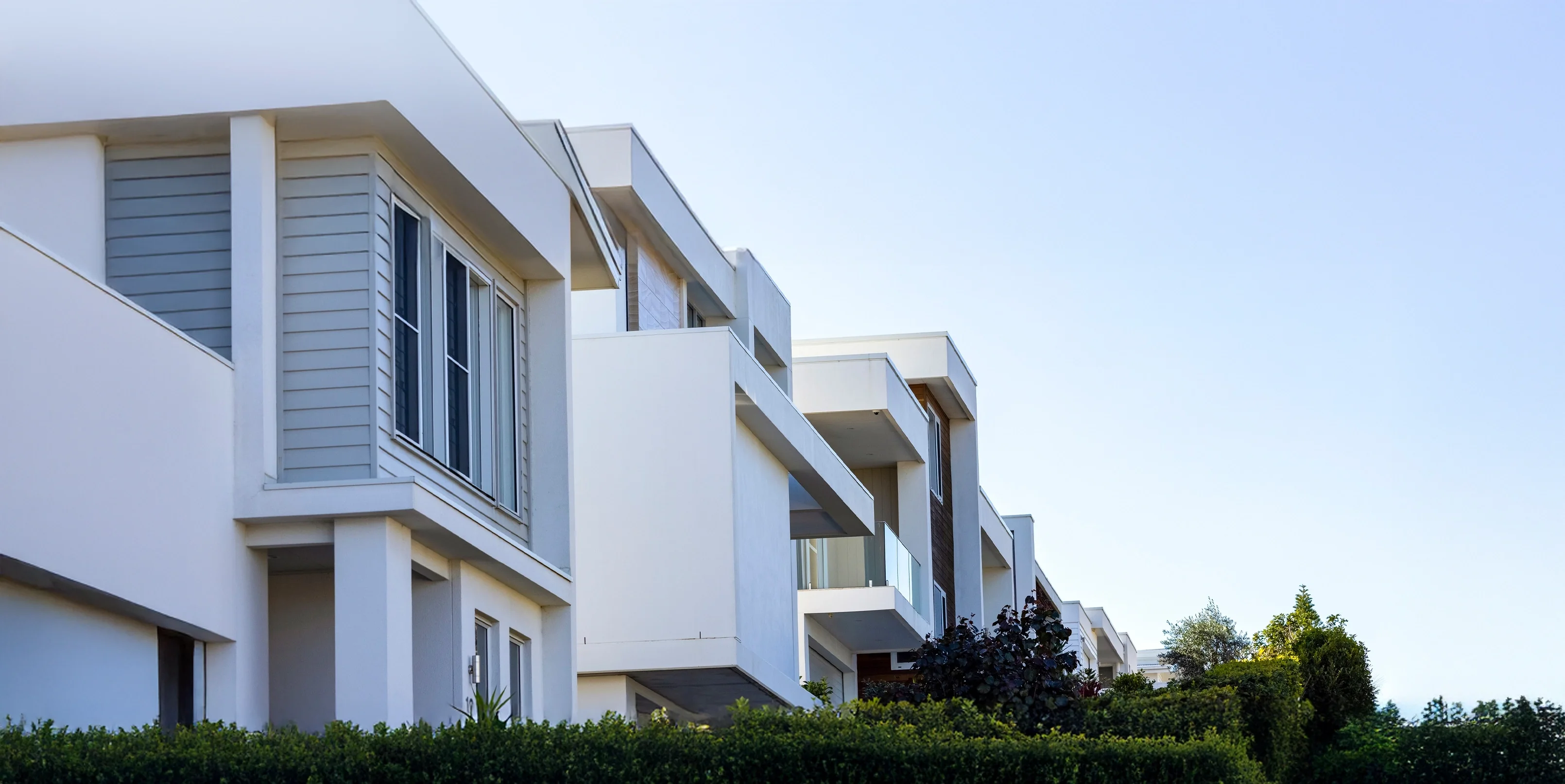 Home & Land HQ Logo branding over Modern white and grey townhouses with  balcony shutters and  trimmed trees.