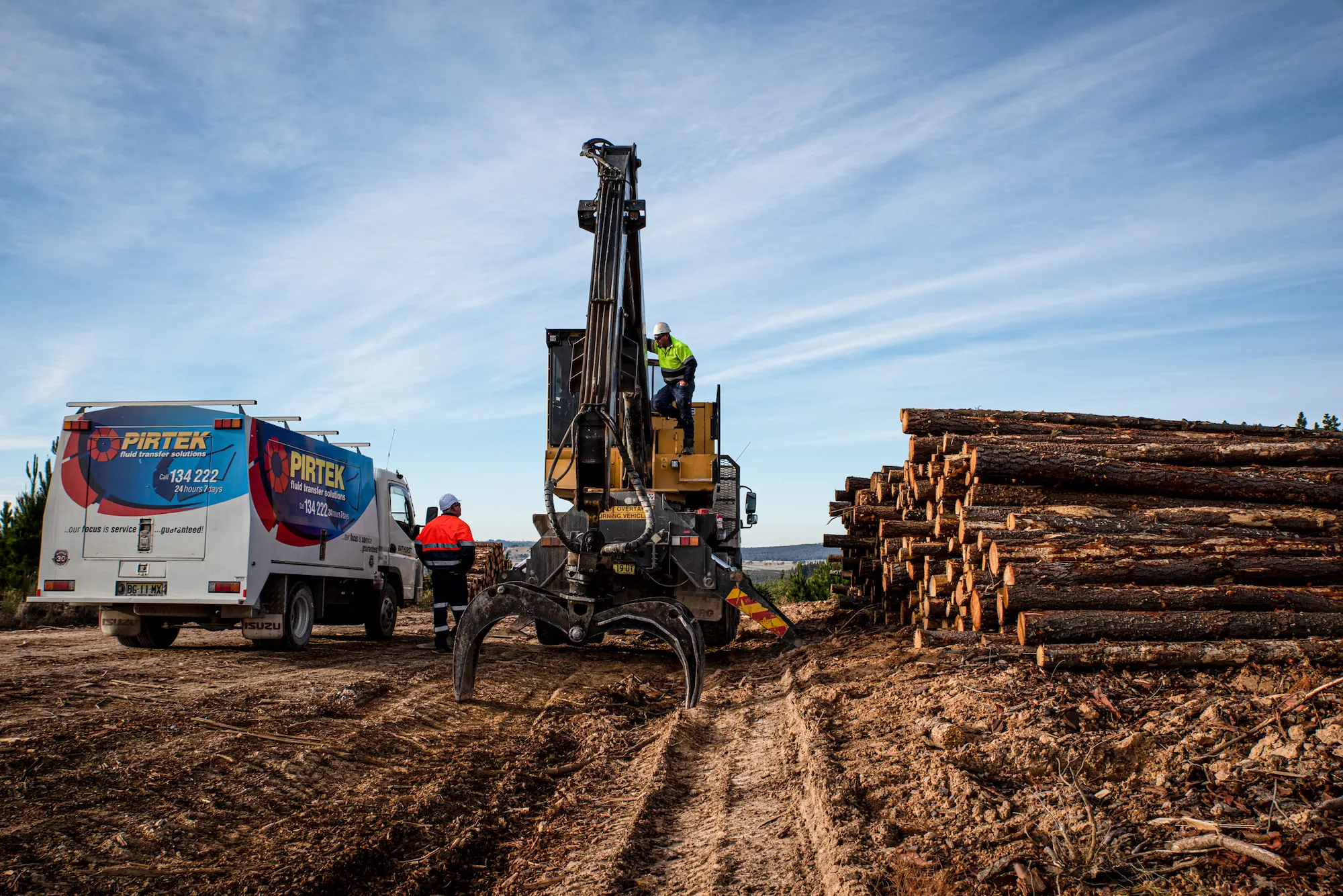 A Pirtek mobile service truck providing onsite support at a forestry location, pictured alongside heavy logging machinery and a large stack of timber under a blue sky.