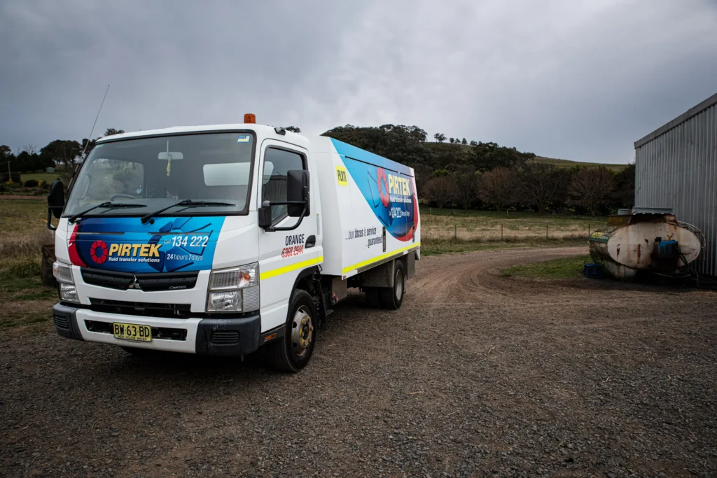 A Mitsubishi Canter service truck displaying full Pirtek livery and contact details for the Orange branch, parked on a gravel area next to a corrugated shed and rural farmland.