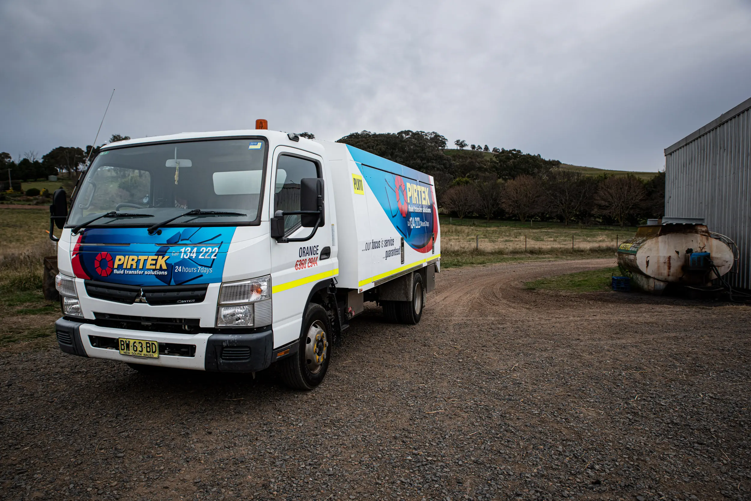 A Mitsubishi Canter service truck displaying full Pirtek livery and contact details for the Orange branch, parked on a gravel area next to a corrugated shed and rural farmland.