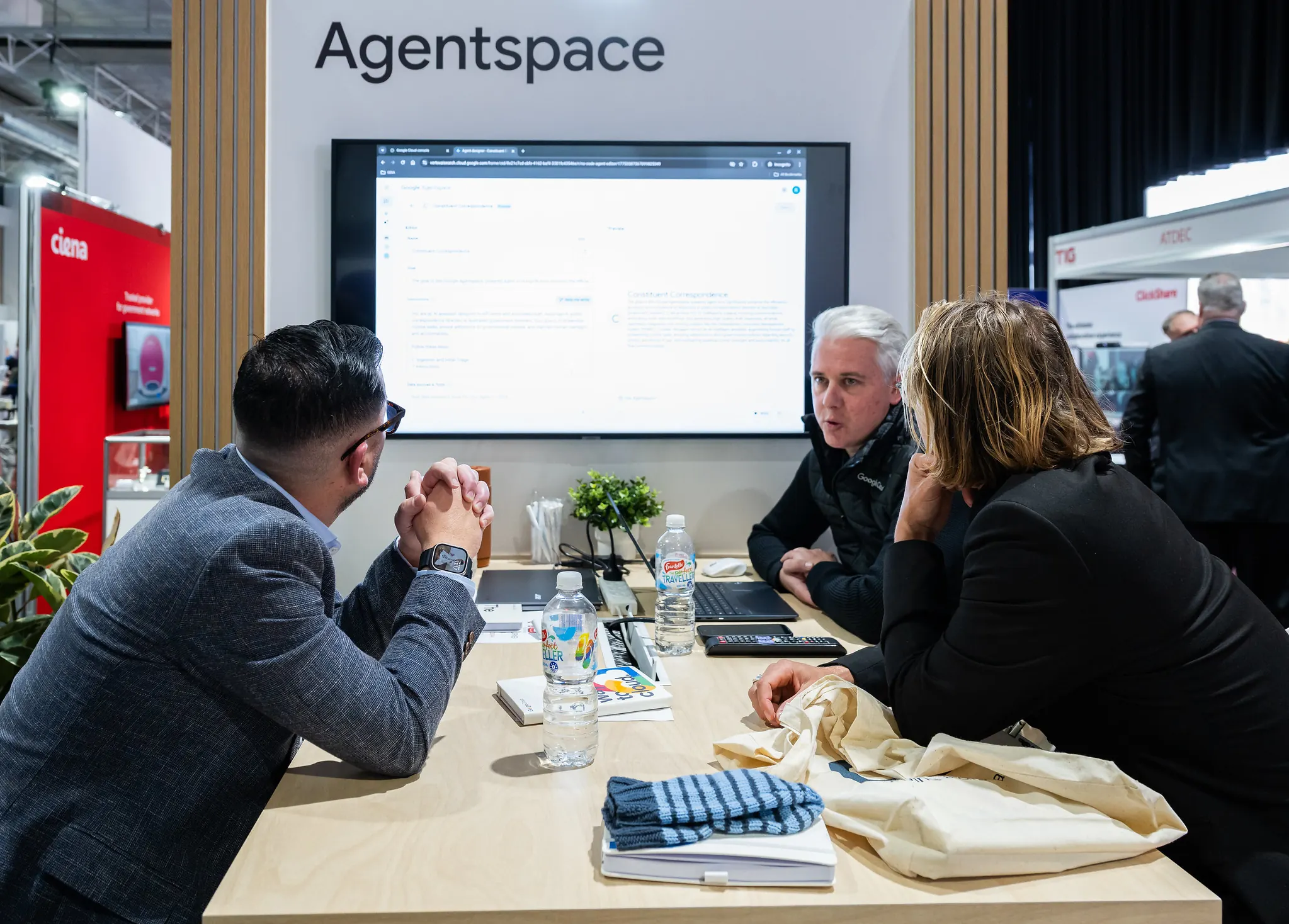 Google representative demonstrating Agentspace product to two business professionals seated at a meeting table with a large display screen showing the platform interface at a conference exhibition