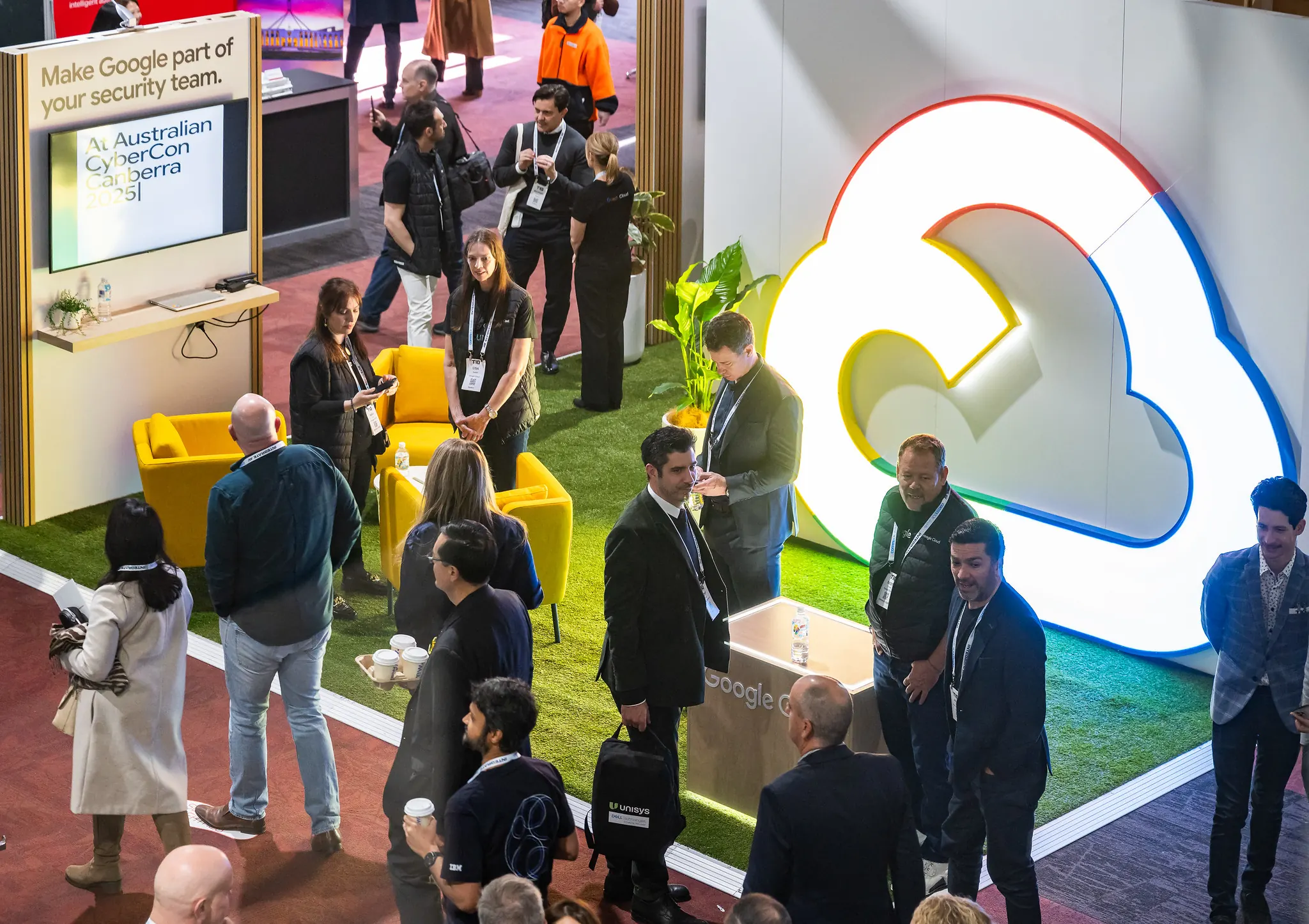 Aerial view of crowded Google Cloud exhibition stand at Australian CyberCon Canberra 2025 showing the illuminated cloud logo, yellow armchairs, artificial turf flooring, and attendees networking with staff
