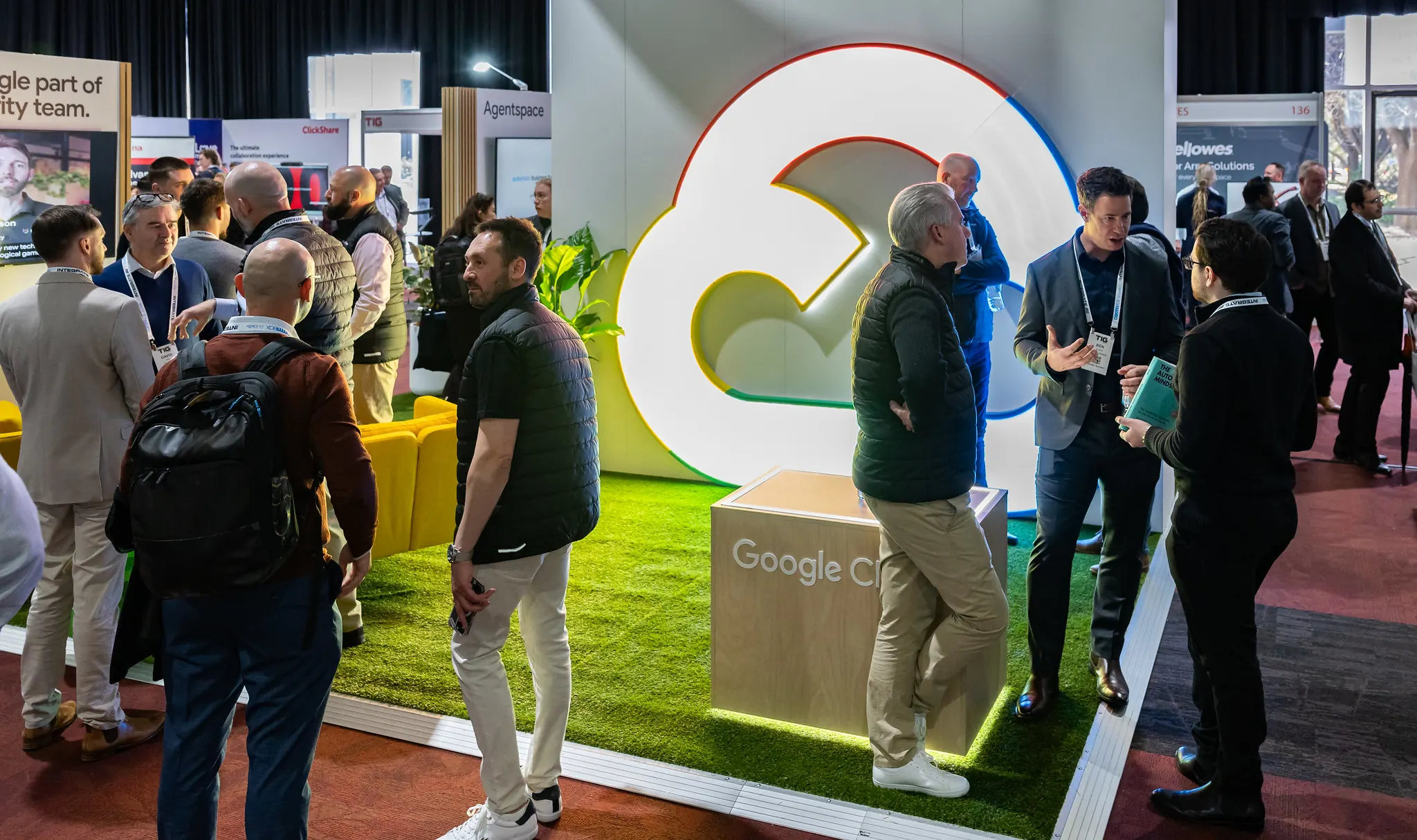 Attendees networking at the Google Cloud exhibition stand featuring a large illuminated Google Cloud logo, artificial grass flooring, and yellow lounge seating at a busy technology trade show